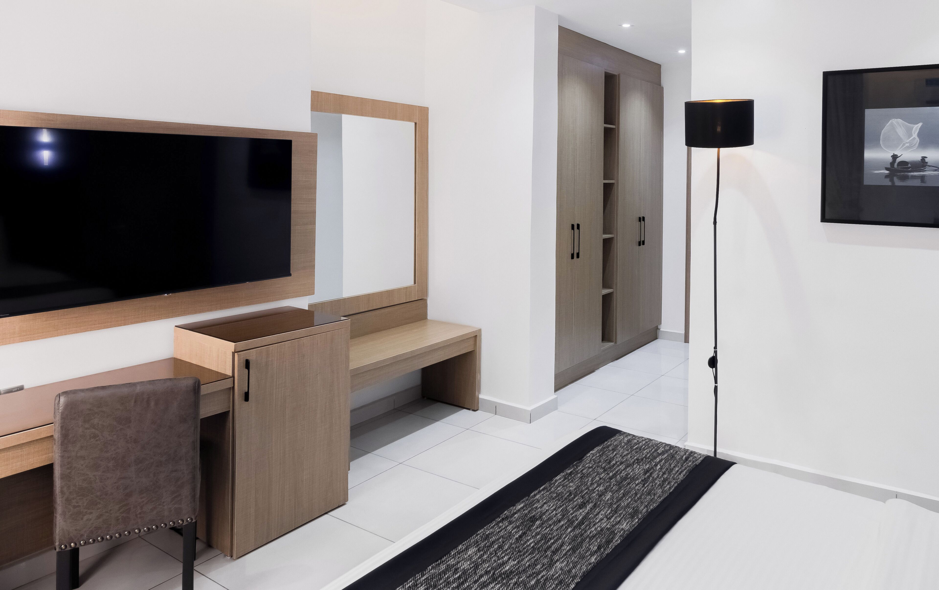 A modern hotel room with a large flat-screen TV, a wooden desk with a chair, a wardrobe, a floor lamp, and a framed picture on the wall.