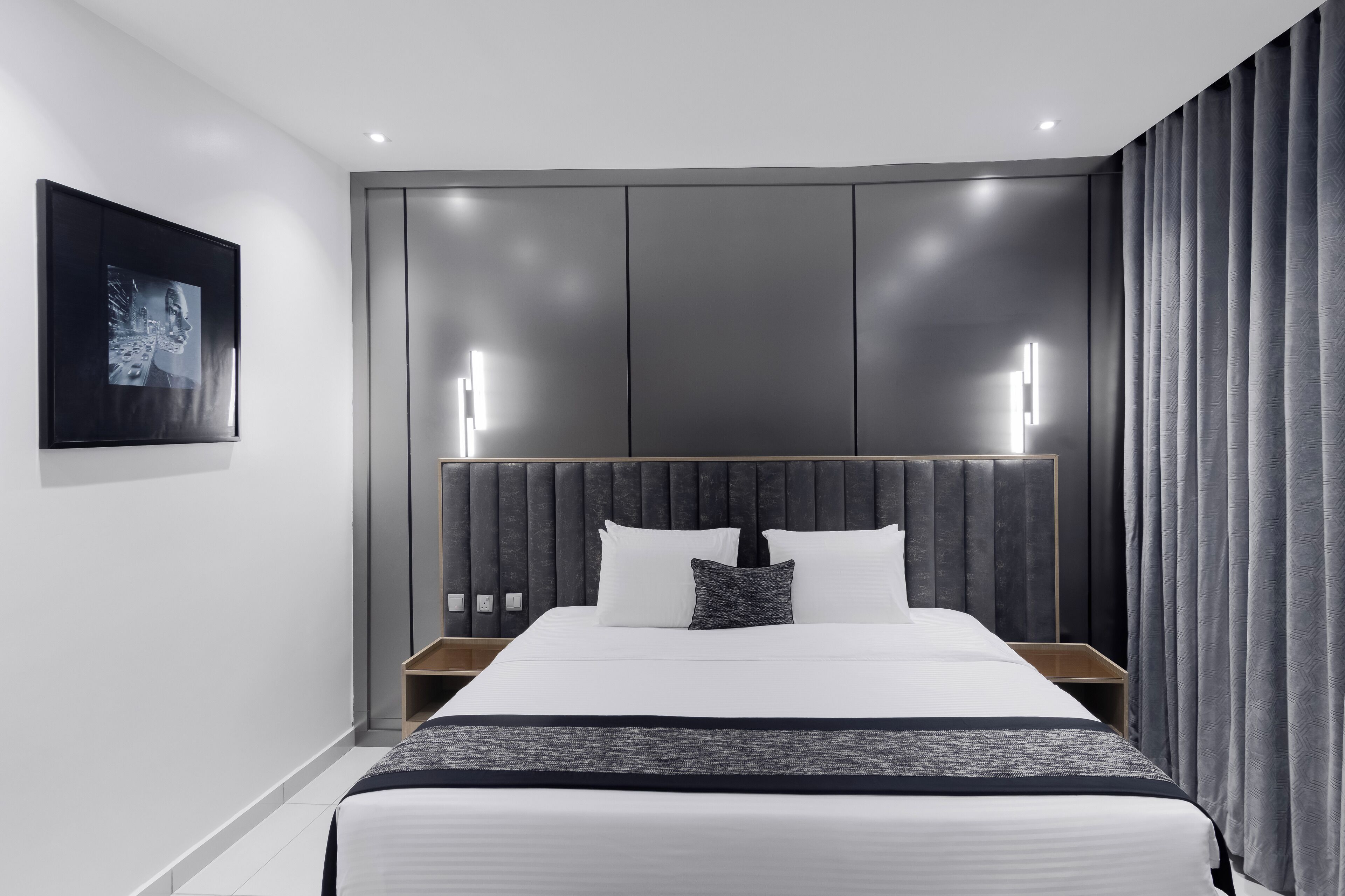 A modern hotel room with a large bed, a headboard with integrated lighting, a bedside table, and a framed picture on the wall.