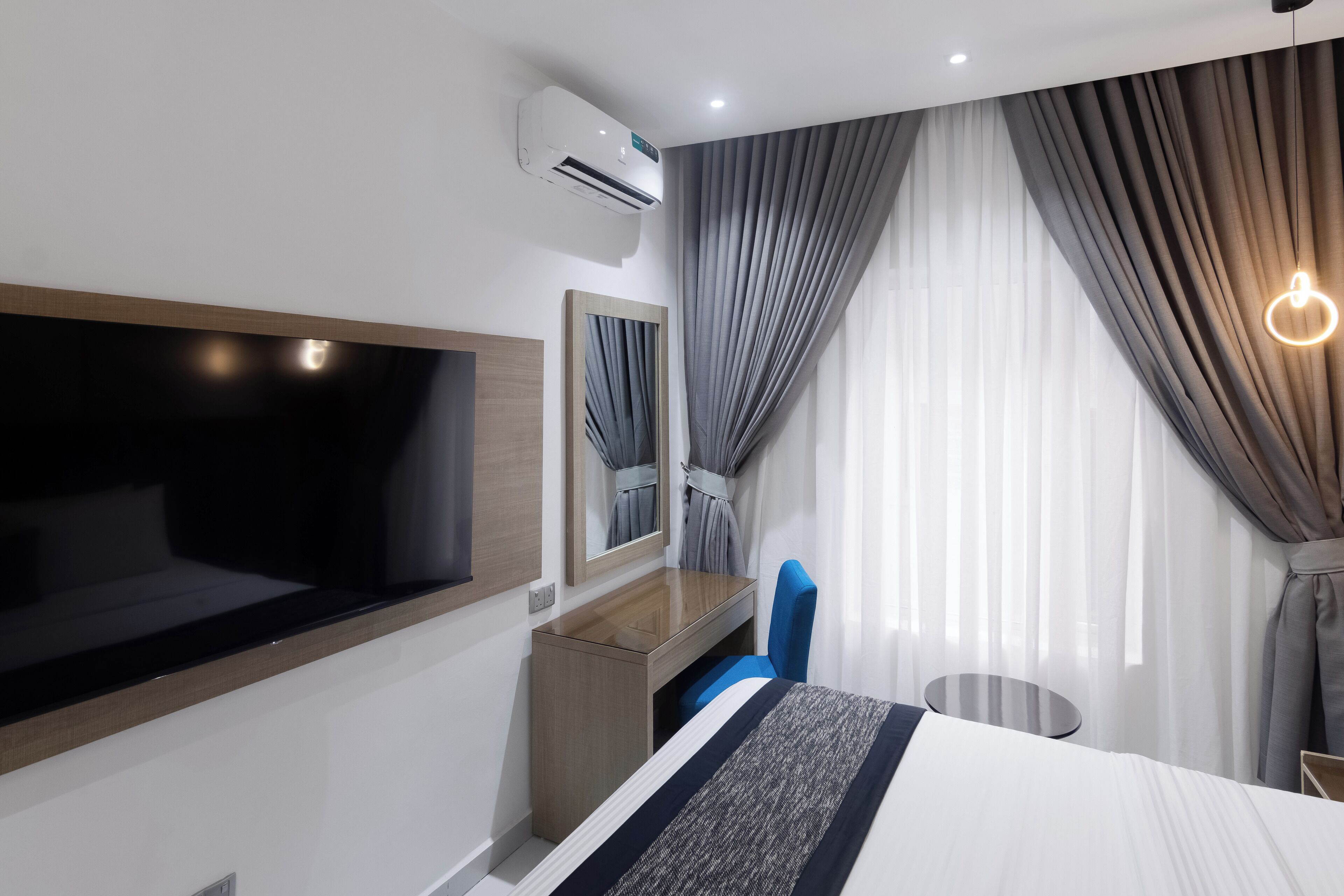A hotel room with a bed, a desk, a chair, a TV, a mirror, and an air conditioning unit.