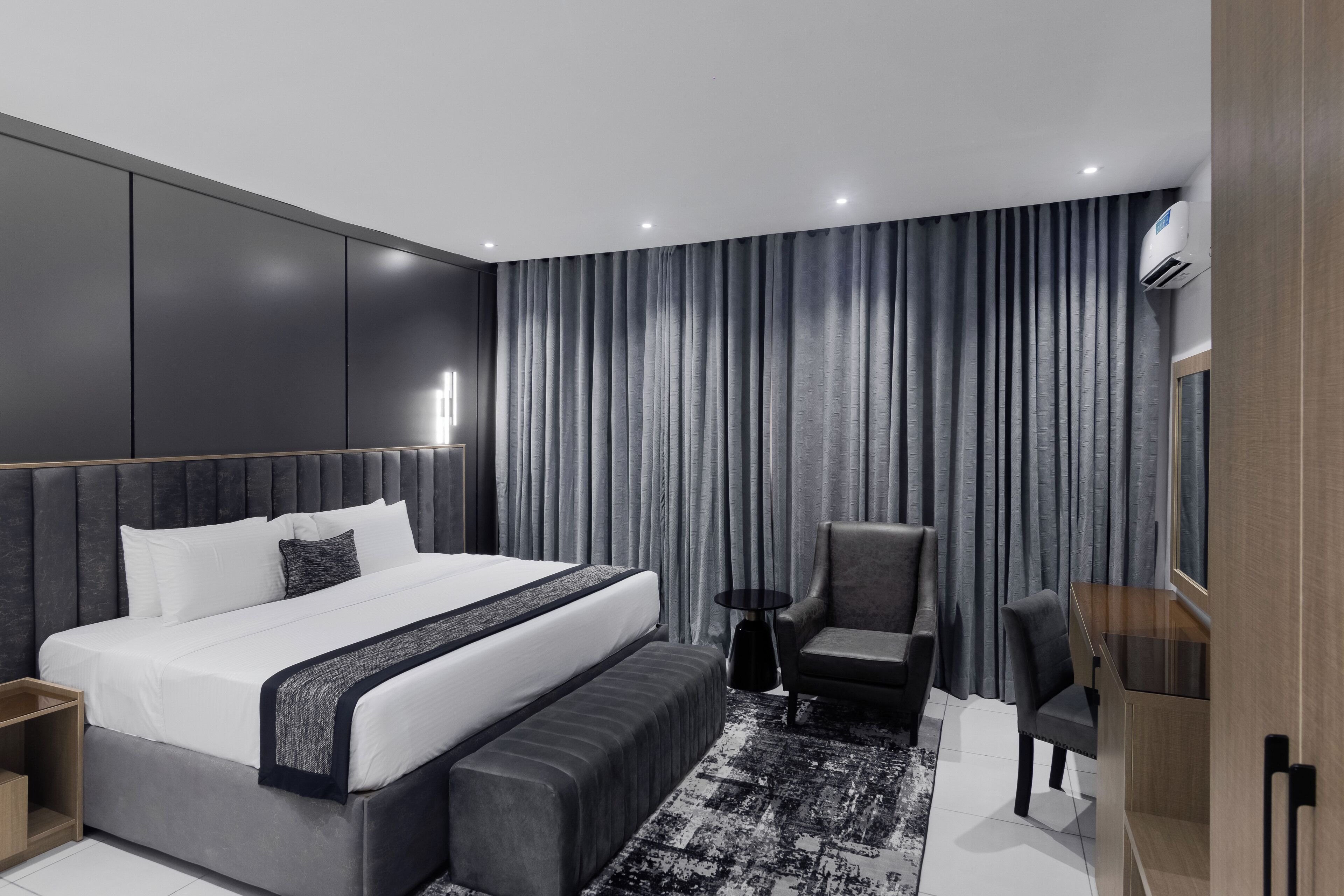 A modern hotel room with a large bed, a side table, a chair, a small round table, and an air conditioning unit.