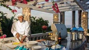 Uno chef prepara il cibo in una cucina esterna con vista sull'oceano, una griglia e un'area pranzo con piatti e tovaglioli.