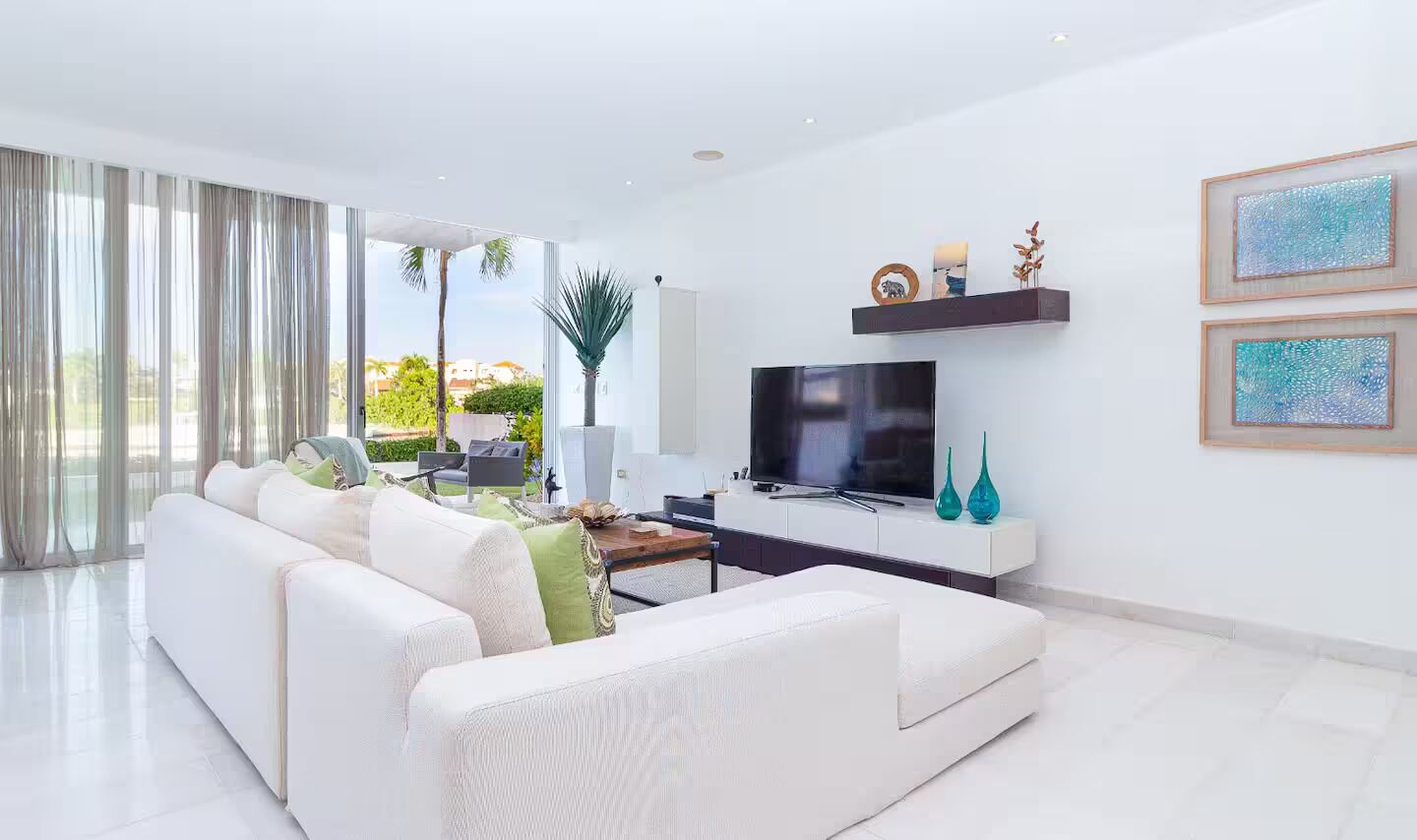 A modern living room with a white sofa, a flat-screen TV, and a large window with curtains.