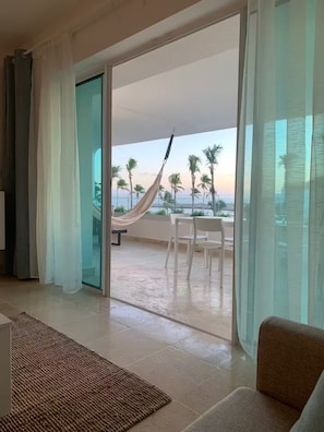 Ein Zimmer mit Blick auf Palmen und das Meer, eine Hängematte sowie ein Essbereich mit Stühlen.