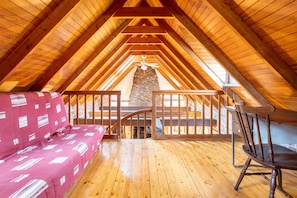 Une chambre confortable située sous les toits, avec un plafond en bois incliné, une cheminée en pierre, un banc en bois et un canapé rouge à motifs.