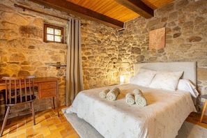Une chambre aux murs de pierre, avec un bureau en bois, un lit sur lequel sont pliés des serviettes et une petite fenêtre.