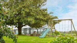 A playground with a slide and swings, a tree, and a greenhouse in the background. - Breton Character House with Shared Pool (Ergué-Gabéric)