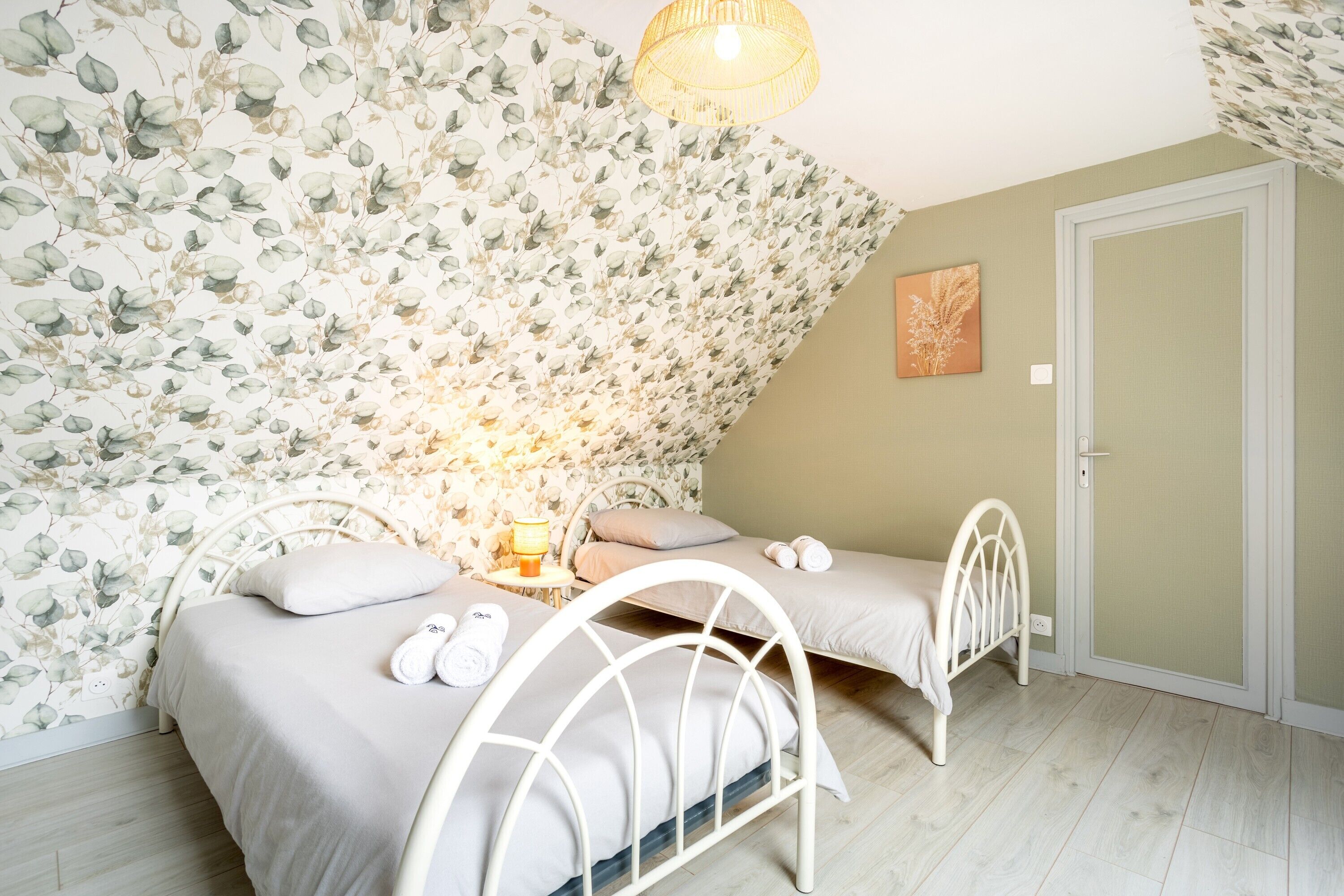 Un dormitorio con papel tapiz floral, dos camas, una mesita de noche con lámpara y una puerta.