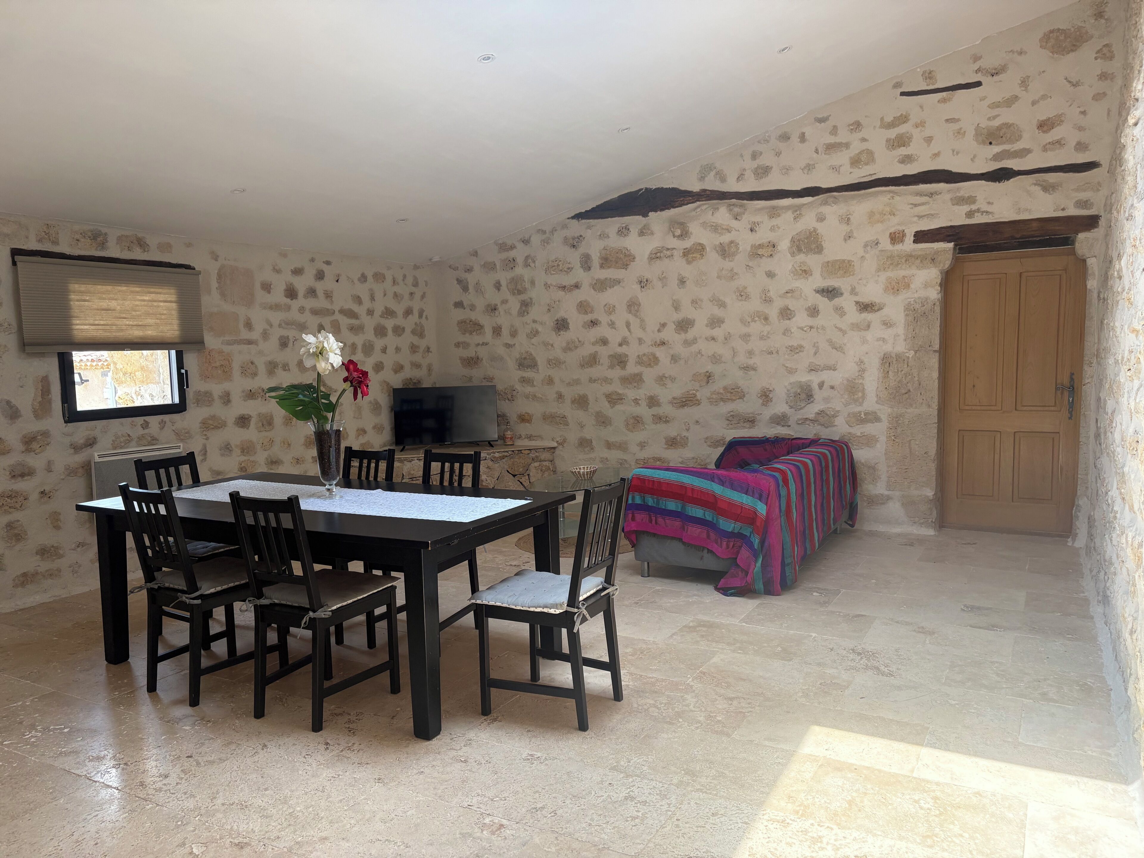 Una habitación de paredes de piedra con una mesa de comedor, sillas, un televisor y una cama con una manta colorida.