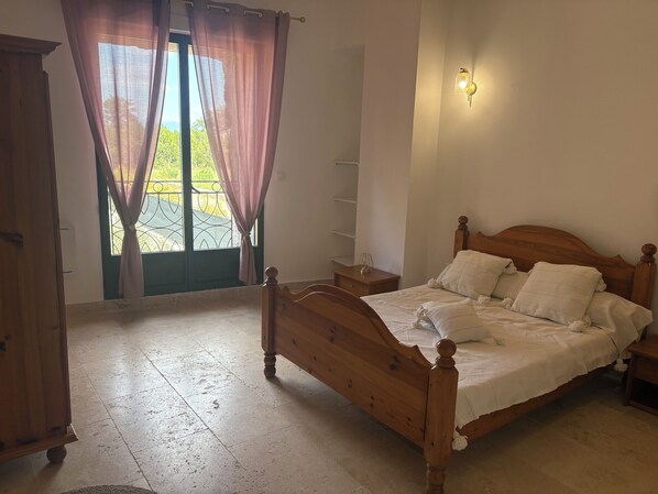 A bedroom with a wooden bed, a nightstand, a window with curtains, and a wall-mounted light fixture. - T3, renovated in a charming estate surrounded by nature in the Haut Var (Régusse)