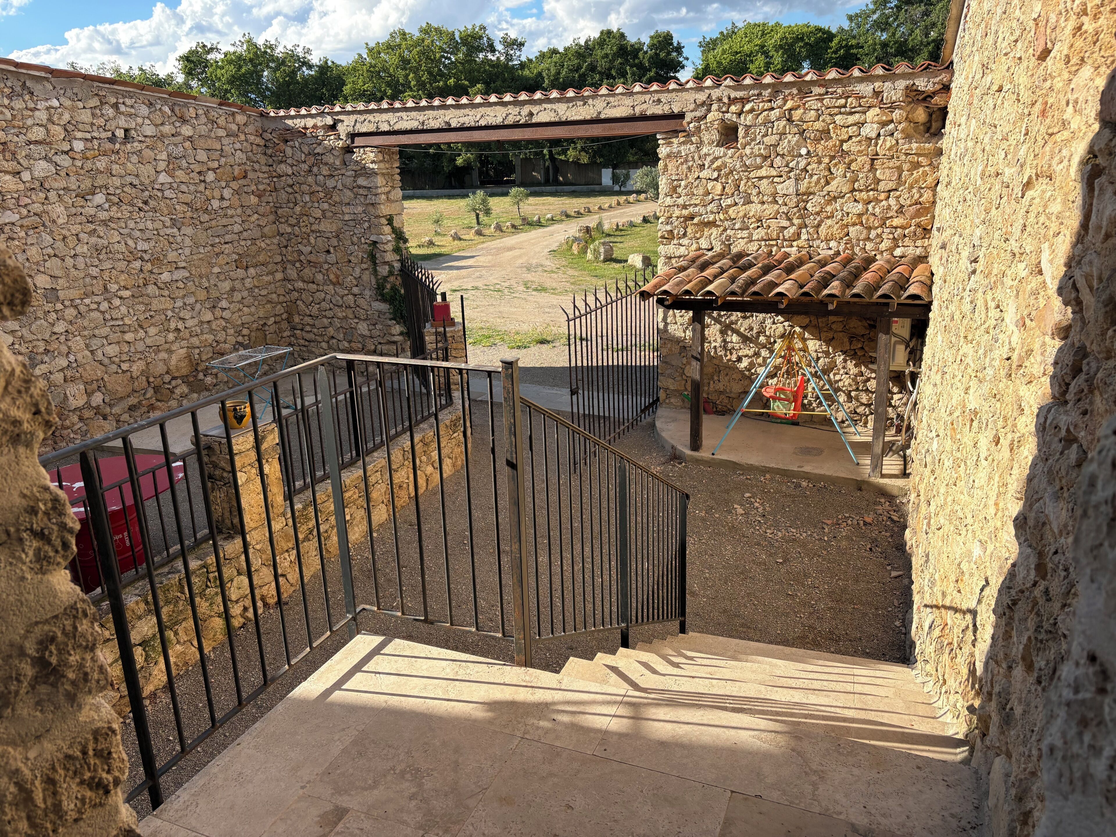 Un bâtiment en pierre avec un portail en métal, un objet rouge sur le sol et un chemin menant à un pont.