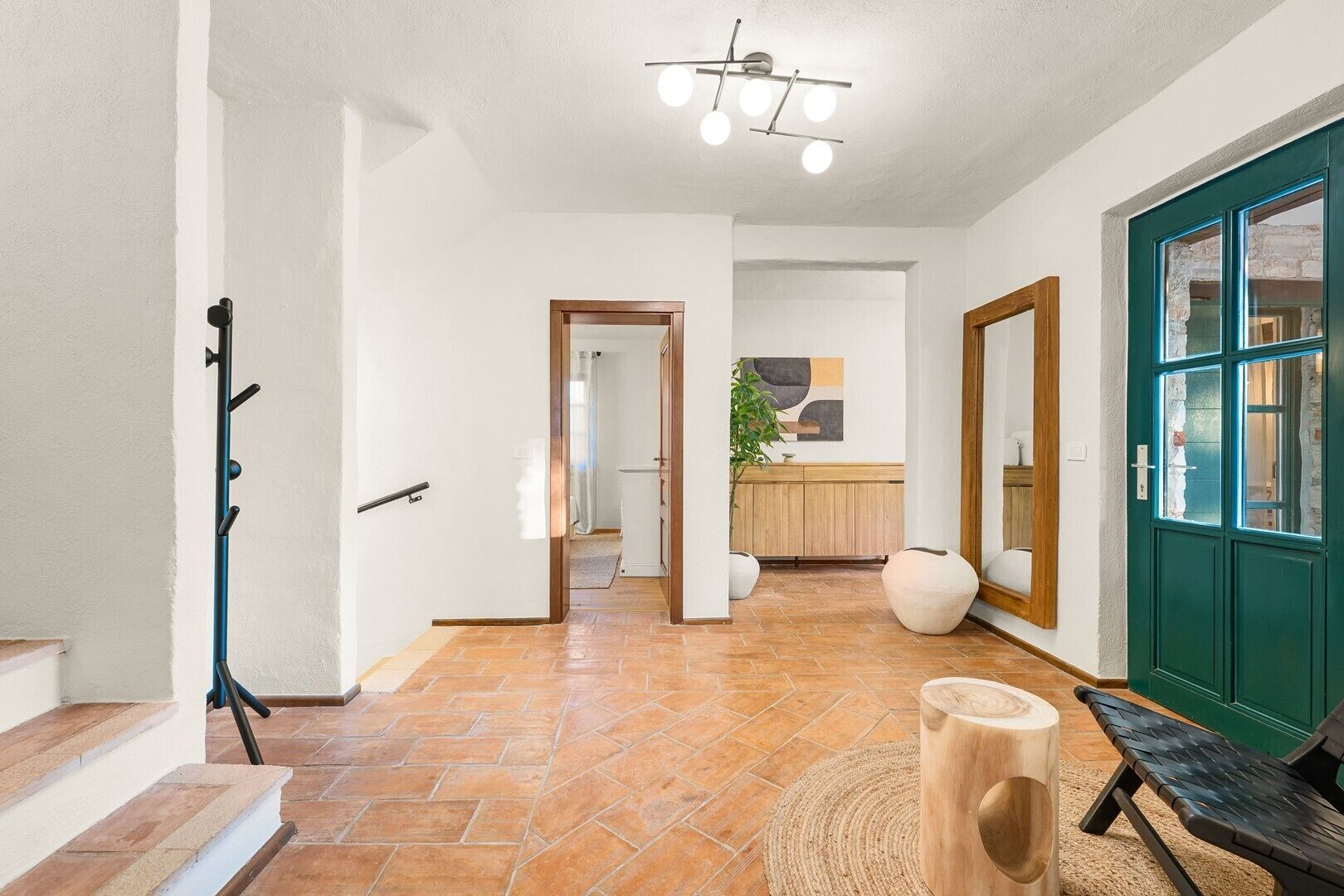 A hallway with a coat rack, a wooden bench, a large vase, and a green door.
