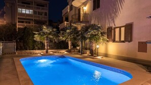 A swimming pool with a curved edge, surrounded by palm trees and a building with balconies. - Charming apartment with pool and parking in Crikvenica (Crikvenica)