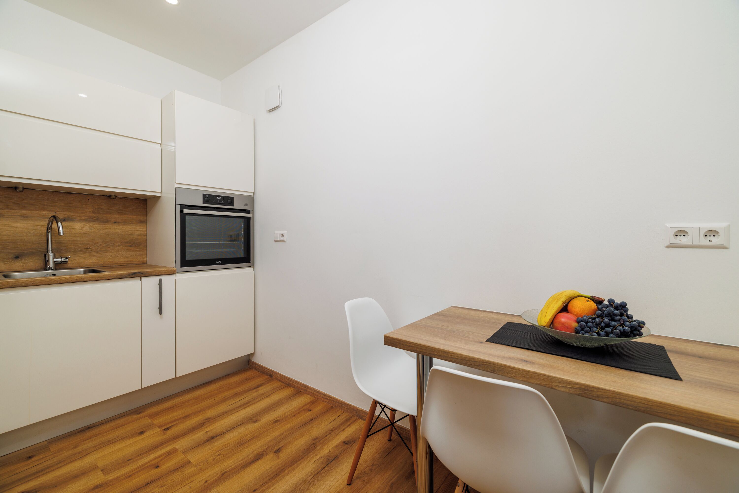 Una cocina moderna con armarios blancos, encimera de madera y horno empotrado. Hay una mesa de comedor con un frutero y sillas blancas.