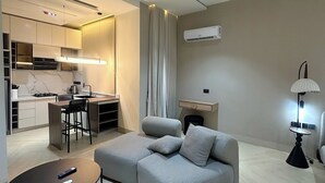 A modern living room with a grey sofa, a round coffee table, a dining area with a kitchen in the background, and a lamp on the right. - Lia's Court Apartment (Abuja)