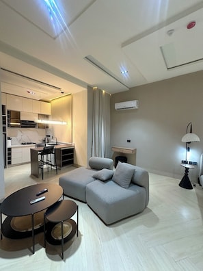 A modern living room with a grey sofa, a round coffee table, a dining area with a kitchen in the background, and a lamp on the right. - Lia's Court Apartment (Abuja)