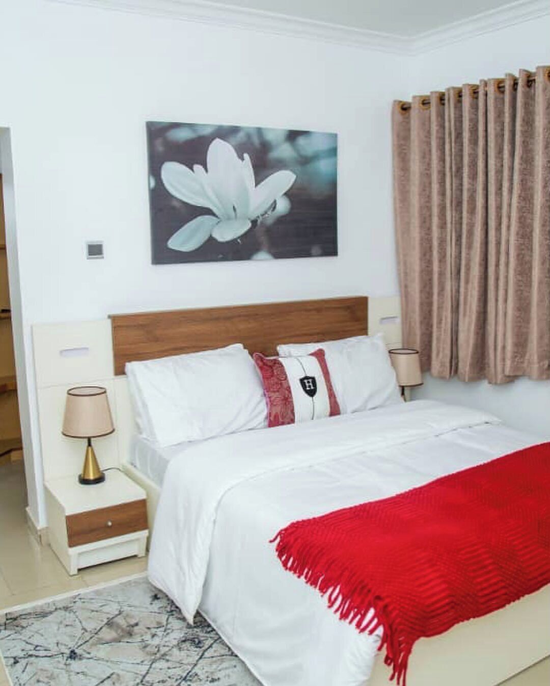 Een slaapkamer met een houten hoofdbord, wit beddengoed, een rode plaid, een nachtkastje met een lamp, en een lijst met een bloem aan de muur.