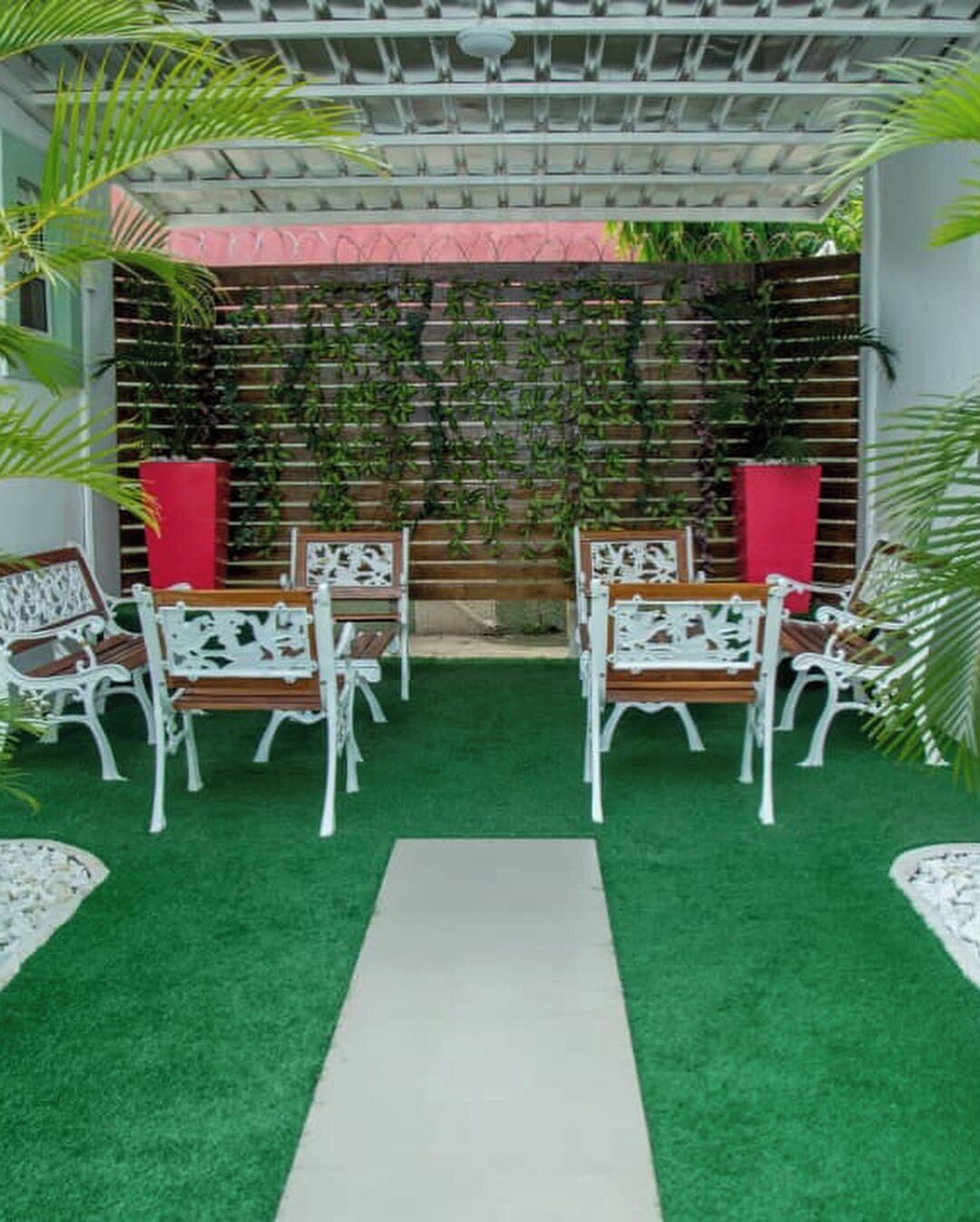 A courtyard with artificial green turf, white ornate chairs, a central pathway, and a backdrop of climbing plants and a wooden fence.