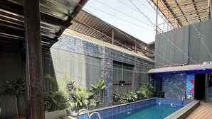 A swimming pool with a tiled edge, surrounded by a wooden deck, under a metal roof structure. - Lia's Court Apartment (Abuja)