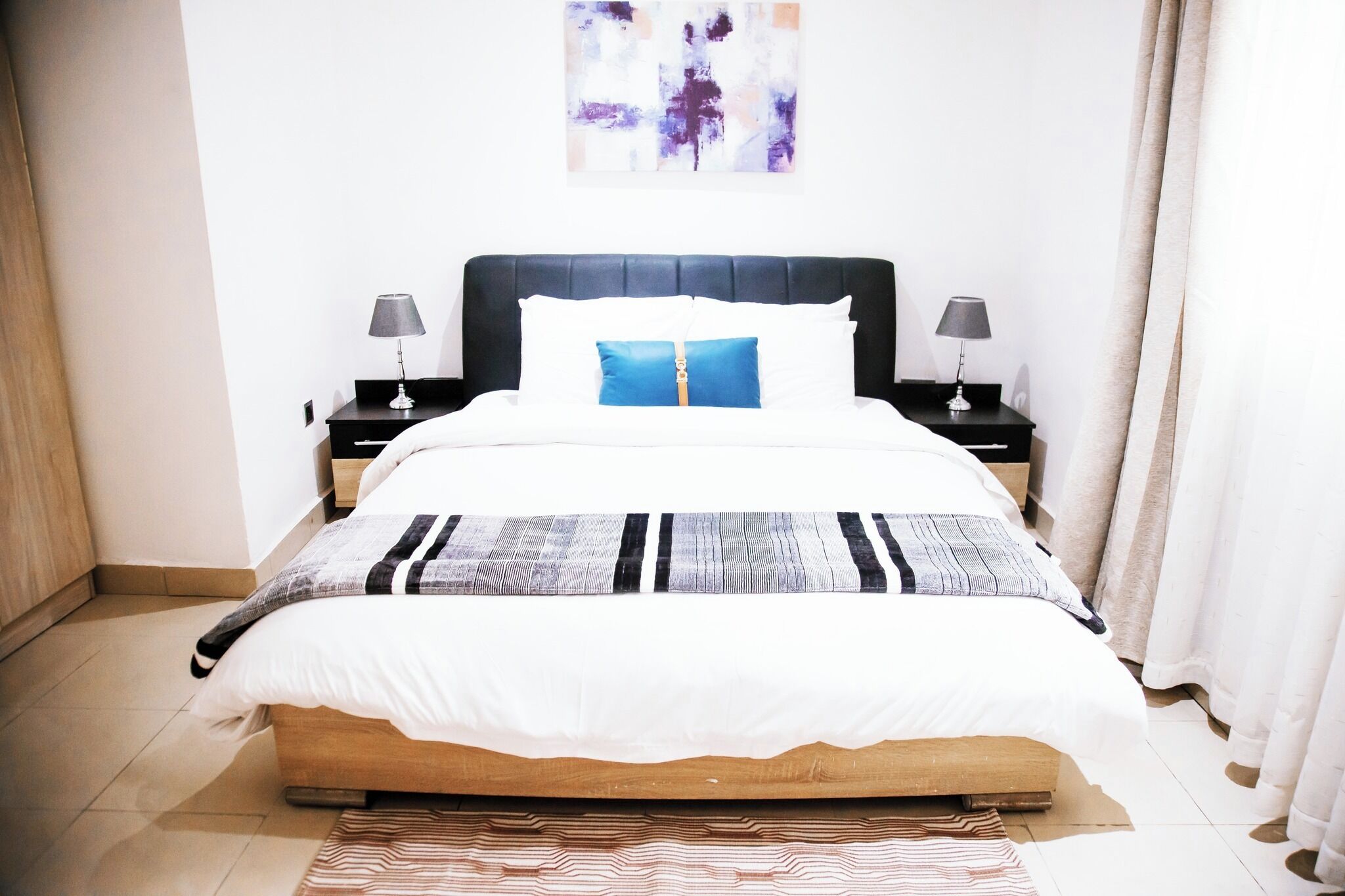 A neatly made bed with a blue pillow, two bedside tables with lamps, a wooden headboard, and a plain wall with a painting.