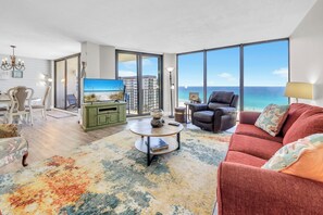 Ein Wohnzimmer mit Sofa, Sesseln und Couchtisch, ein Essbereich mit Stühlen und Tisch sowie große Fenster mit Blick auf das Meer.
