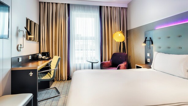 A hotel room with a bed, a desk, a chair, a small table, and a lamp.