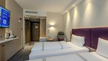 Quarto de hotel com duas camas, uma mesa de trabalho, uma cadeira e uma televisão exibindo informações do hotel.