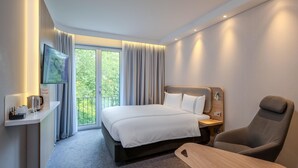 A modern hotel room with a bed, a desk with a coffee maker, a TV, and a view of greenery outside.