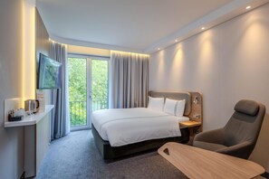 A modern hotel room with a bed, a desk with a coffee maker, a TV, and a view of greenery outside.