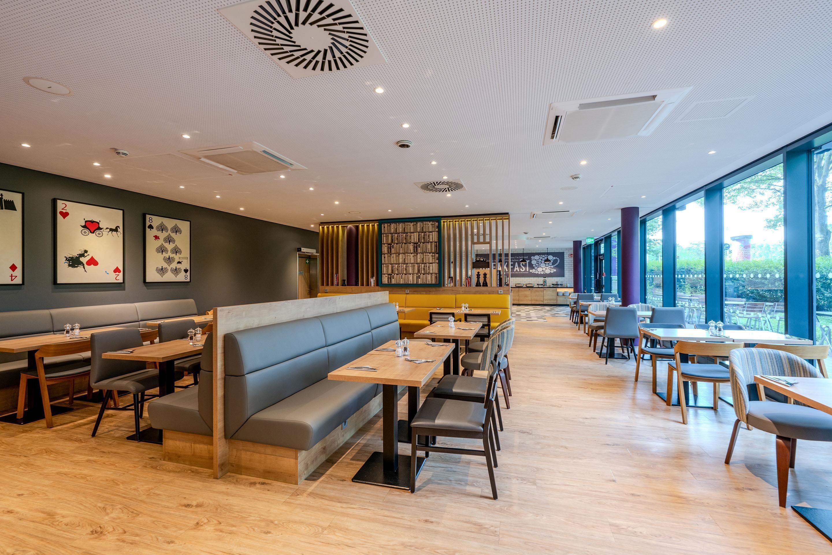 A restaurant with wooden tables, grey and yellow booths, and large windows offering a view.