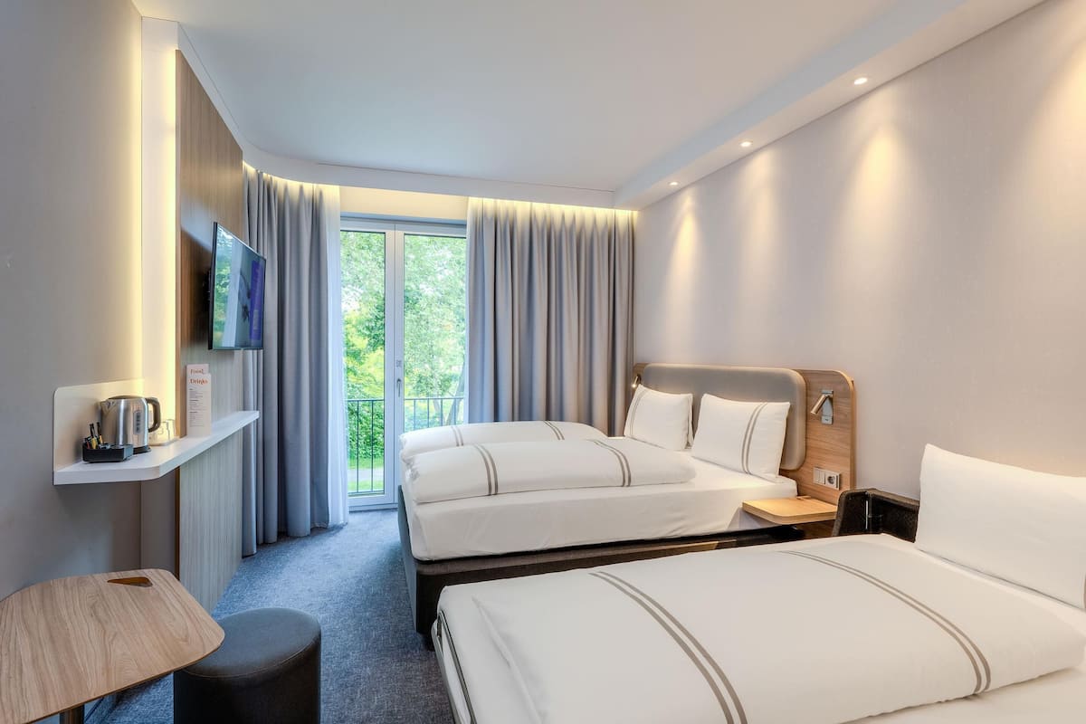 A hotel room with two beds, a TV, a coffee maker, a small table, and a view of greenery through the window.
