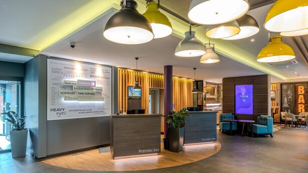 Een moderne receptiebalie van een hotel, met een receptiebalie met de tekst 'Premier Inn', een zithoek met stoelen en een groot scherm dat informatie toont.