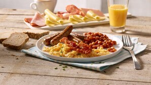 A breakfast plate with eggs, sausages, beans, and toast, a glass of orange juice, and a cup of coffee on a wooden table.