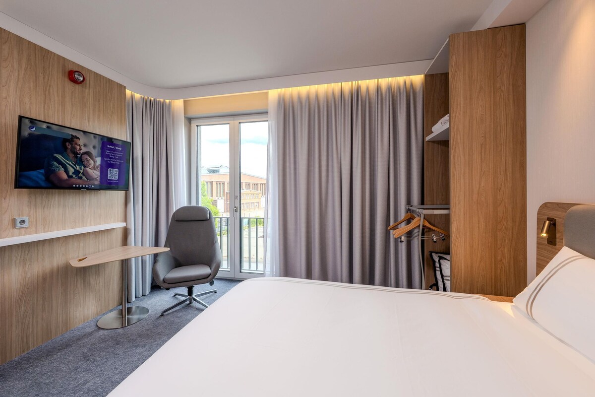 A hotel room with a bed, a desk, a chair, a TV mounted on the wall, and a view of the outside.