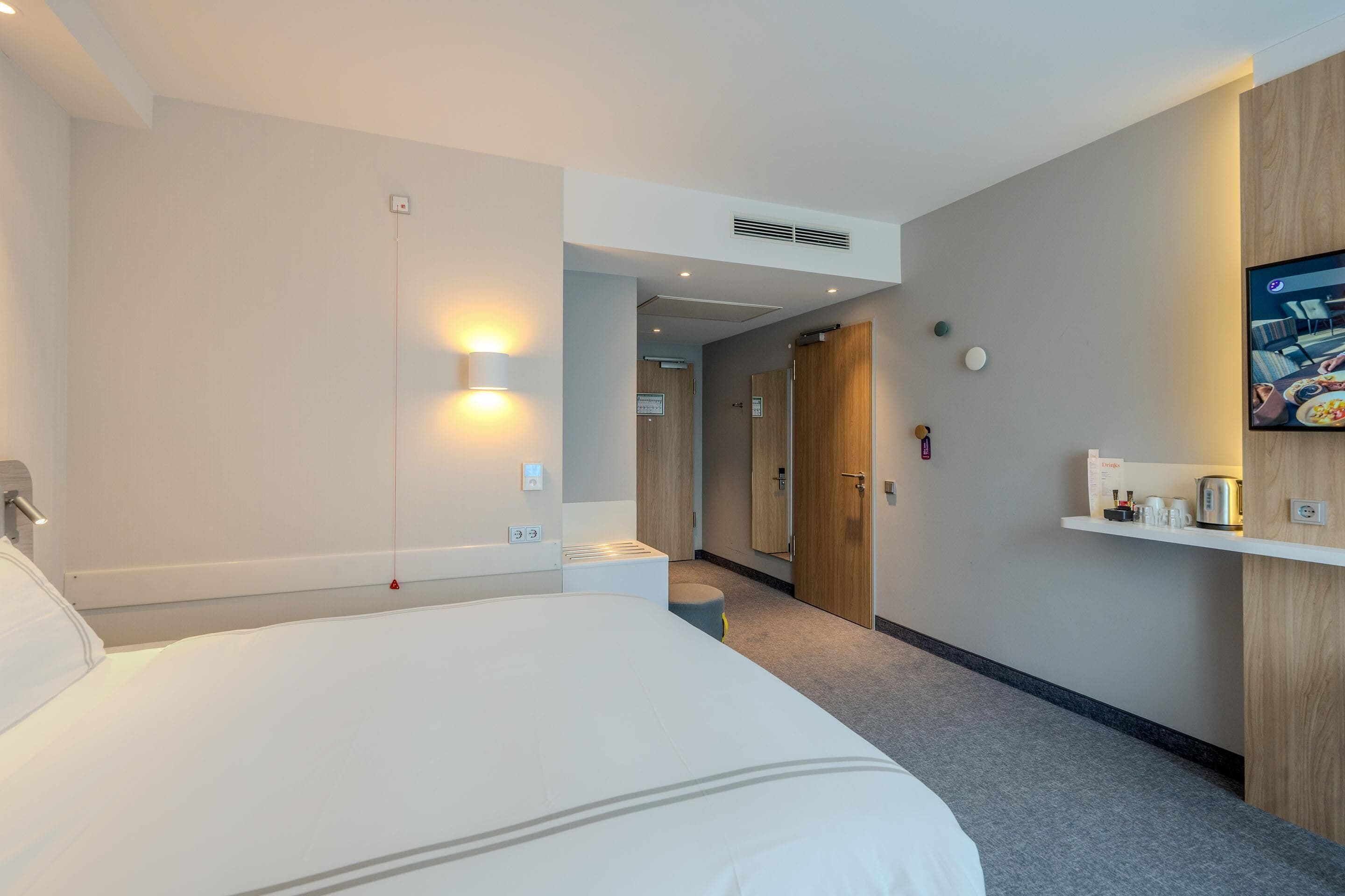 A hotel room with a large bed, a bedside table, a wall-mounted TV, and a small shelf with items.