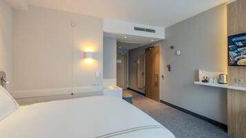 A hotel room with a large bed, a bedside table, a wall-mounted TV, and a small shelf with items.