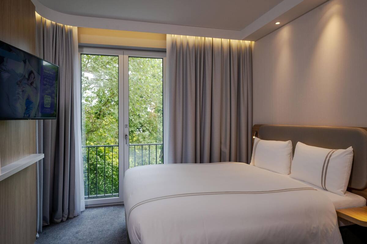 A hotel room with a large bed, a television mounted on the wall, and a balcony with curtains.