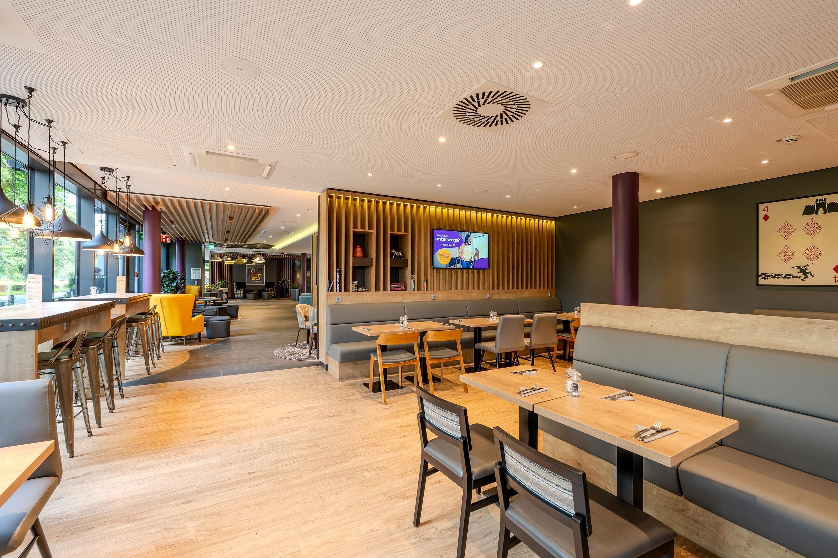 A modern restaurant interior with wooden tables, cushioned seating, and a large TV screen.