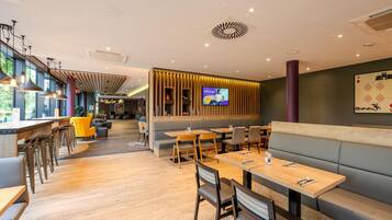 A modern restaurant interior with wooden tables, cushioned seating, and a large TV screen.