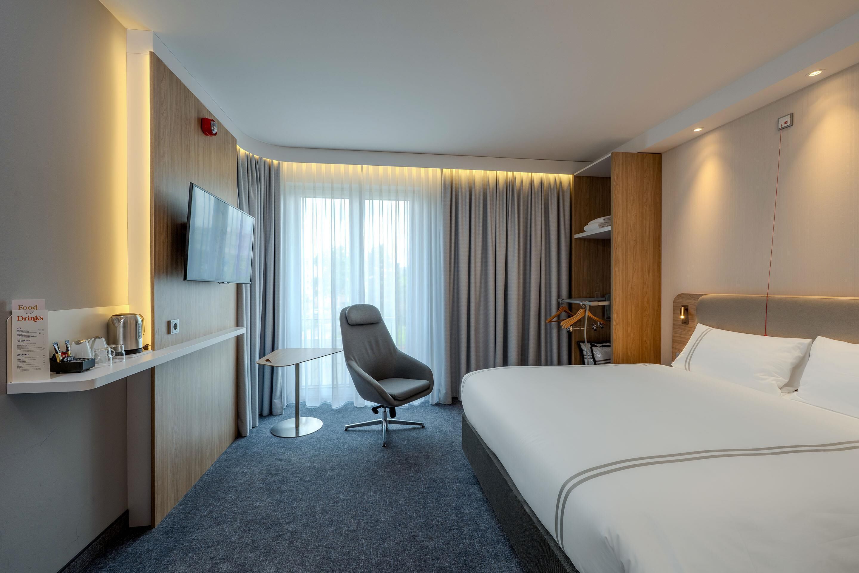 Habitación de hotel con cama, una silla, un televisor, una mesita con hervidor, y un cartel que indica comida y bebidas.