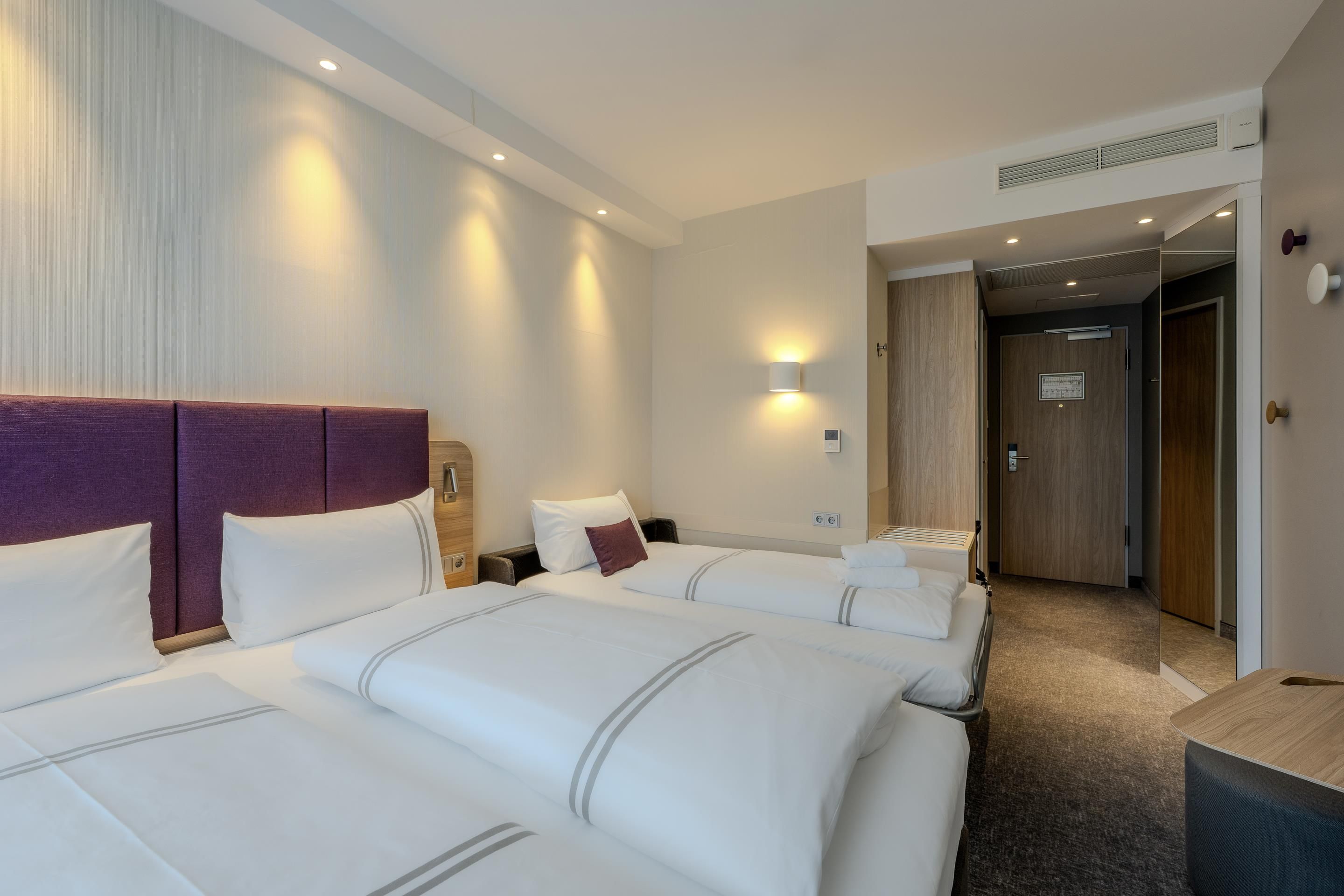 A hotel room with two beds, a wooden headboard, a nightstand, a wall-mounted lamp, and a door leading to another room.