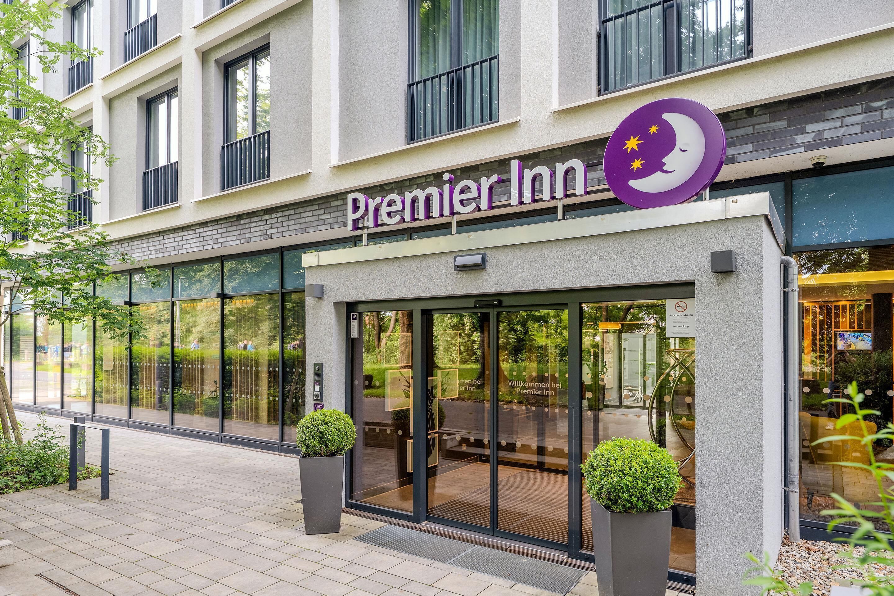 Entrada de un hotel con puerta de cristal, un cartel que dice 'Premier Inn' y dos plantas en macetas afuera.