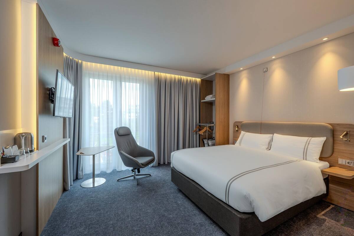 A hotel room with a bed, a desk, a chair, a TV, and a window with curtains.