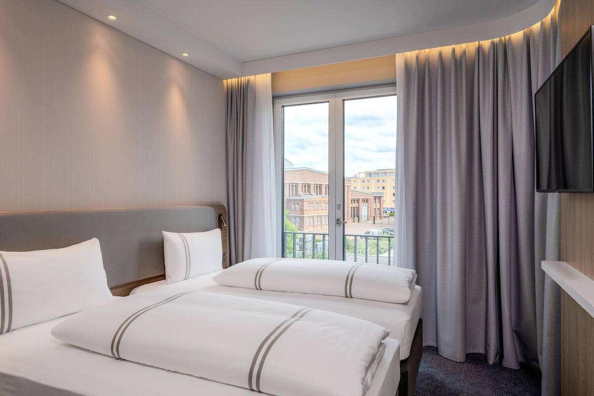 A hotel room with a large bed, a flat-screen TV, a balcony with curtains, and a view of buildings.