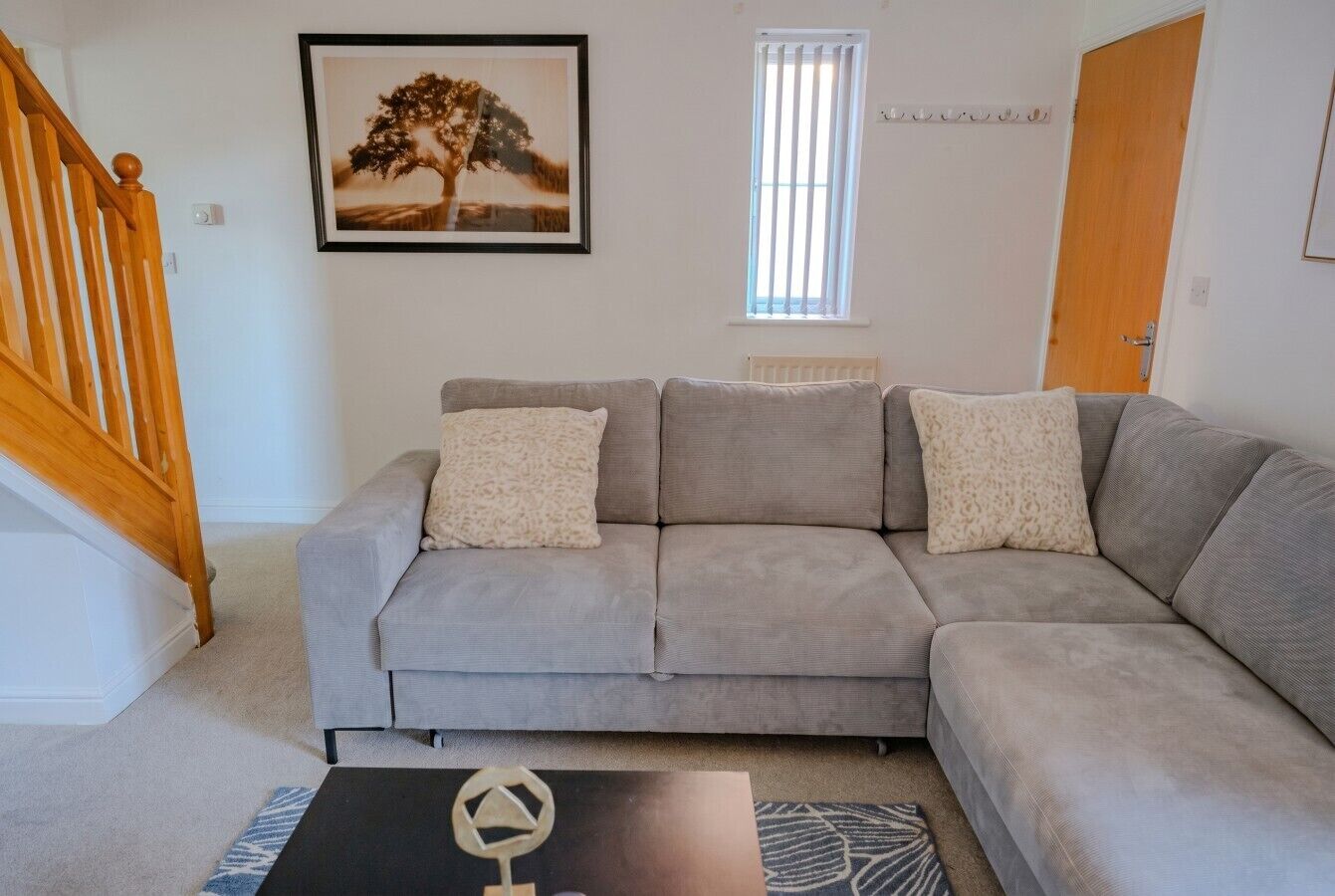 A living room with a grey sofa, two pillows, a black coffee table with a geometric design, a framed picture on the wall, a window with blinds, and a wooden staircase.
