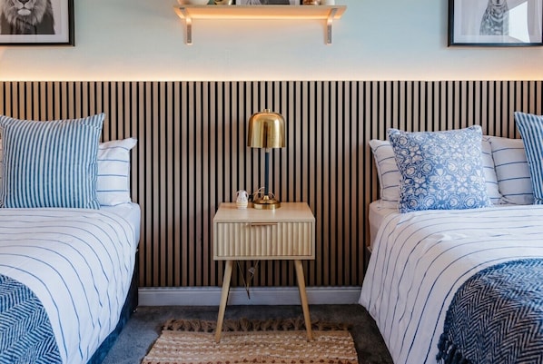 Ein Schlafzimmer mit zwei Betten, einem Nachttisch mit Lampe, einem Wandregal und einem gemusterten Teppich.