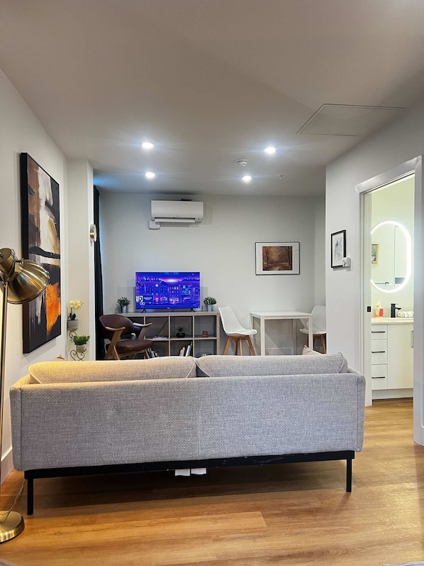 A modern living room with a grey sofa, a flat-screen TV, a desk, and a mirror with lights. - Sunbird Studio, Bright Downtown Escape, AC (Halifax)