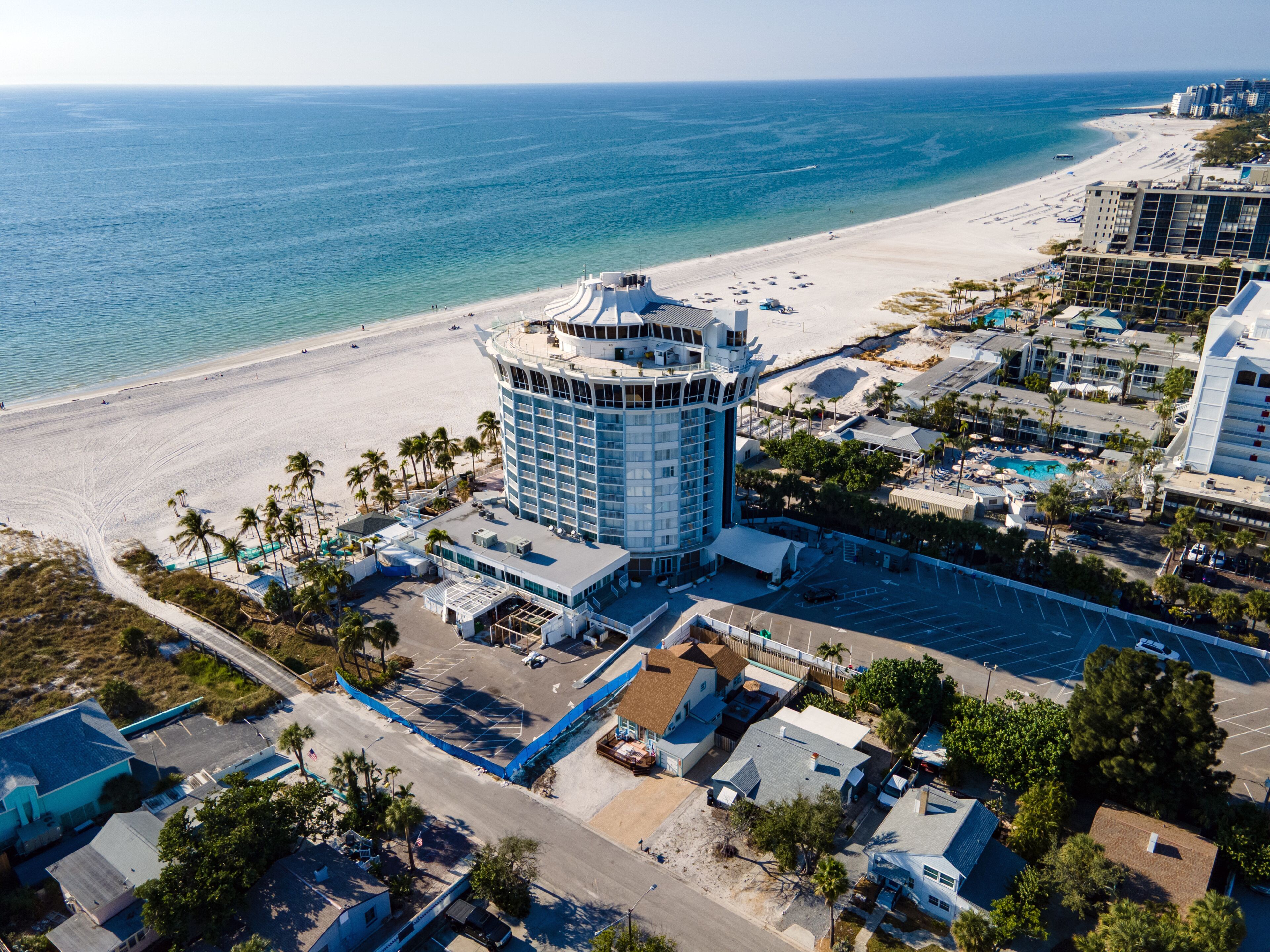 An aerial view of a coastal hotel with a central tower, beachfront, and surrounding buildings.