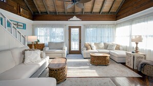 A spacious living room with a high ceiling, wooden beams, a central ceiling fan, and a large door with a window.