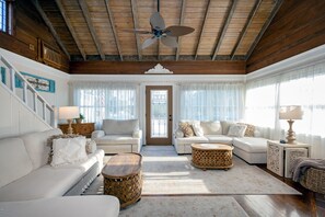 A spacious living room with a high ceiling, wooden beams, a central ceiling fan, and a large door with a window.
