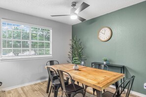 Een eethoek met een houten tafel, zwarte stoelen, een potplant, een wandklok en een plafondventilator.